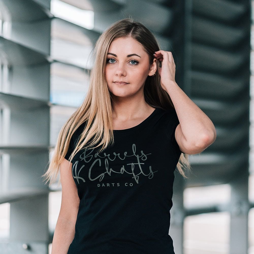 T-shirt damski Barrels and Shafts – czarny Eine Frau trägt ein schwarzes T-Shirt mit der Aufschrift „Barrels and Shafts Darts Co“. Sie steht vor einem modernen, lichtdurchfluteten Hintergrund.