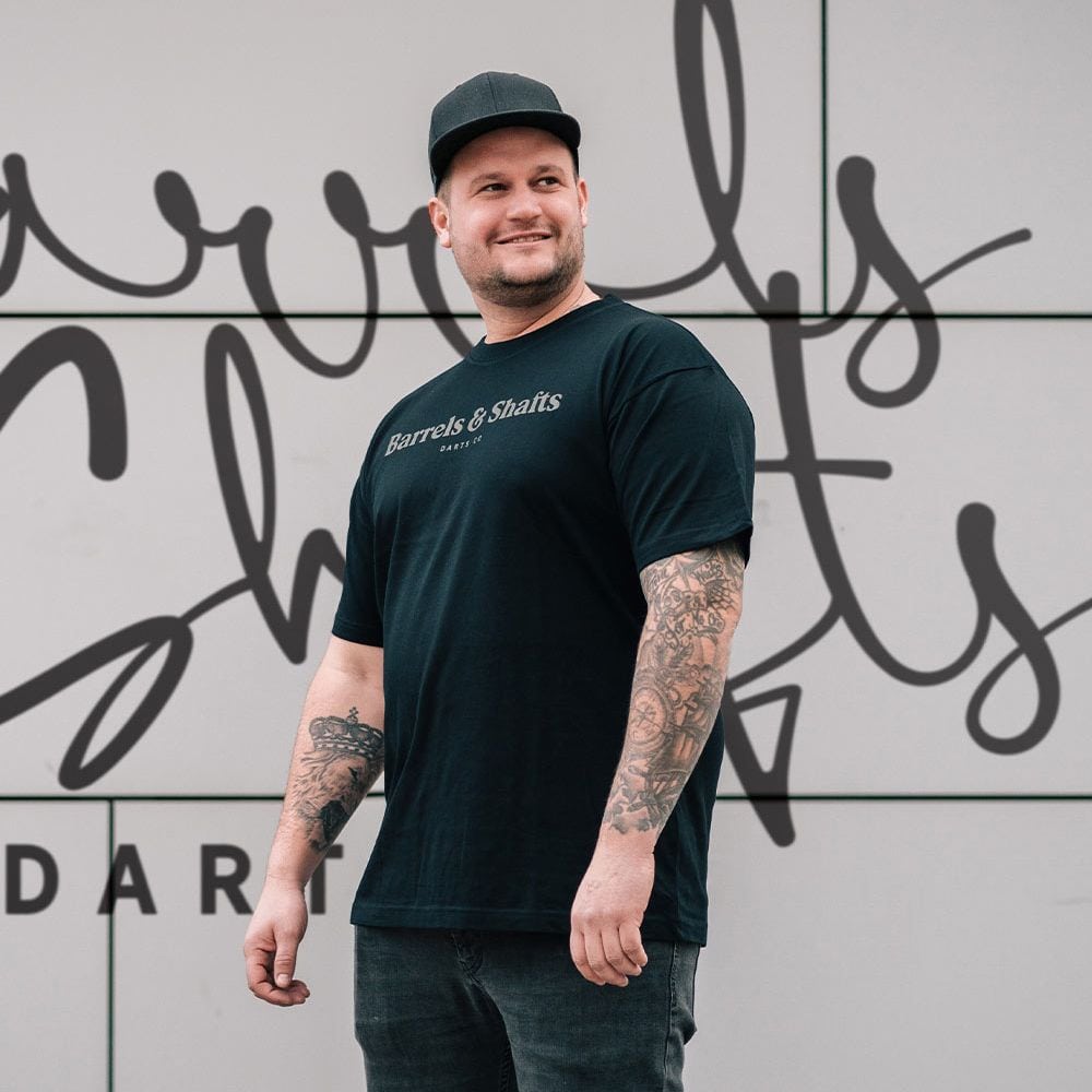 T-shirt „Barrels and Shafts” – czarny Ein Mann trägt ein schwarzes T-Shirt mit der Aufschrift „Barrels & Shafts“. Er steht vor einer Wand mit passender Schrift im Hintergrund.