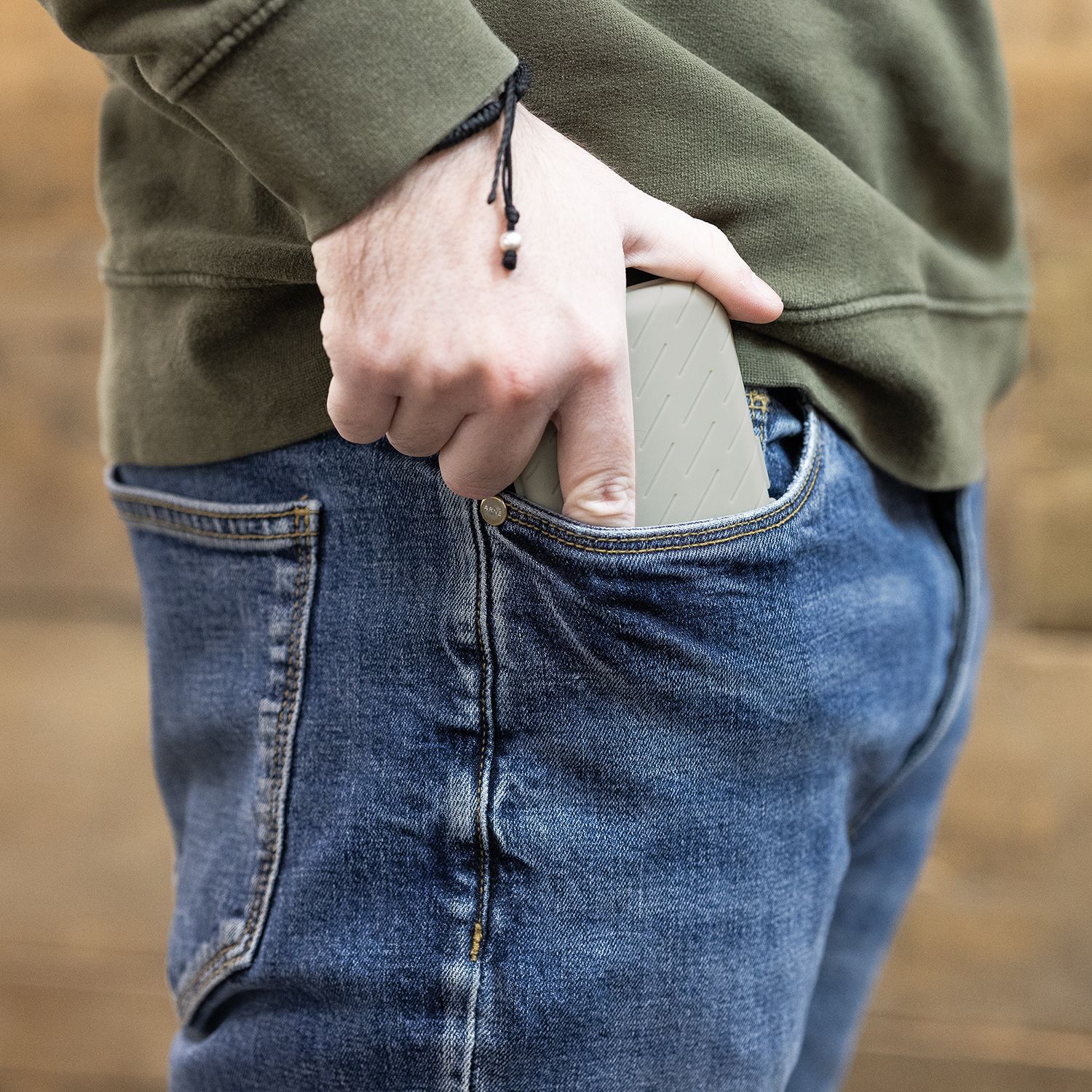 Eine Person steckt eine graue Darttasche in ihre Hosentasche. Die Person trägt eine blaue Jeans und einen olivgrünen Pullover.