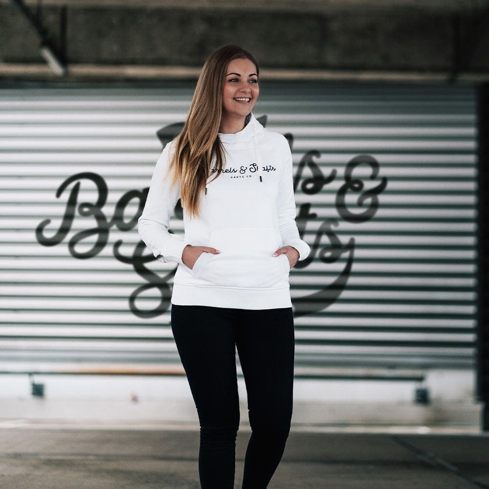 Damska bluza z kapturem Barrels and Shafts – biała Eine Frau trägt einen weißen "Barrels and Shafts" Damen Hoodie. Sie steht vor einem Hintergrund mit großem Schriftzug und lächelt.