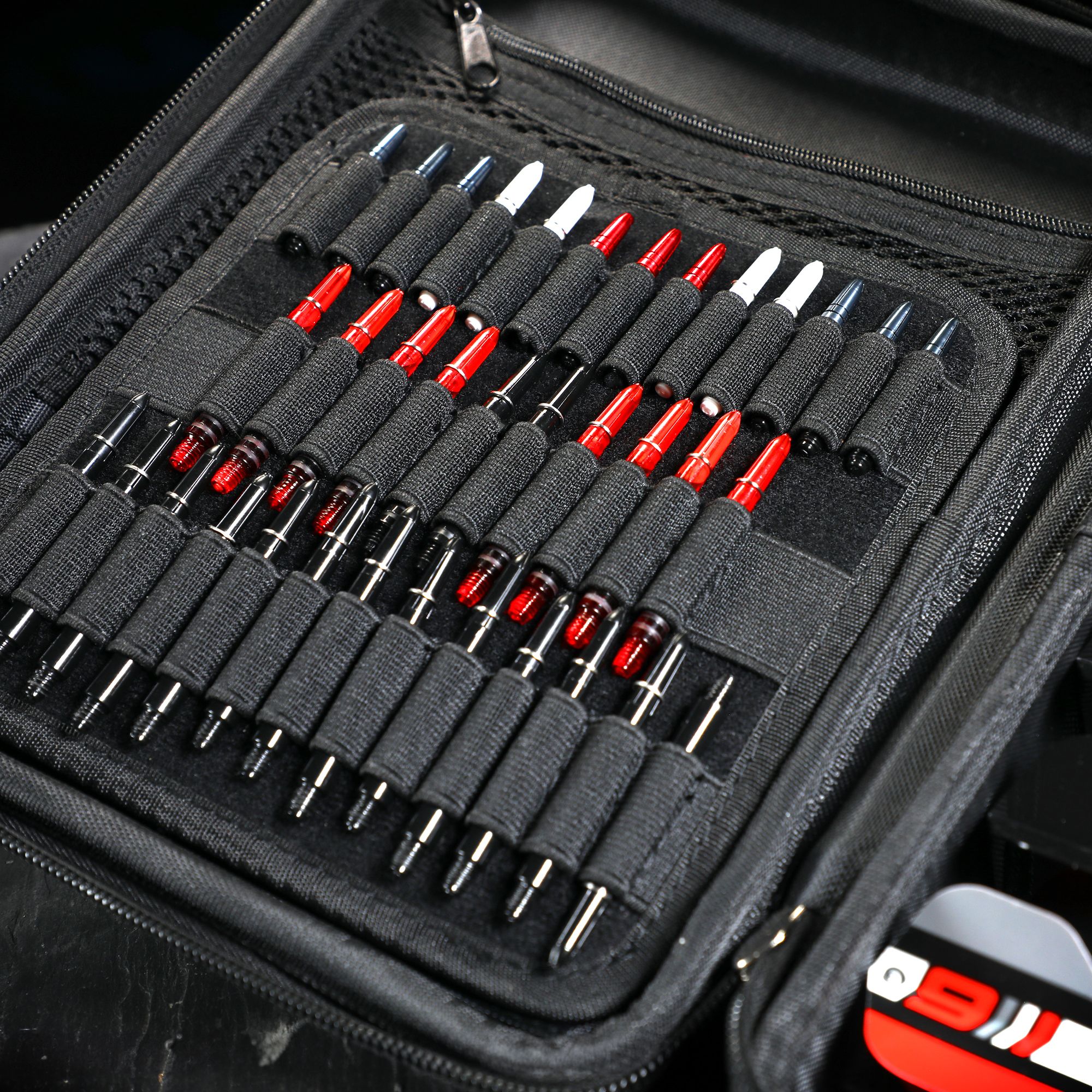 Winmau Armor G4 Dartwallet Das Bild zeigt ein geöffnetes schwarzes Dartwallet mit vielen ordentlich einsortierten Dartspitzen. Die Dartspitzen sind in elastischen Schlaufen gehalten und teilweise rot, weiß oder schwarz gefärbt.