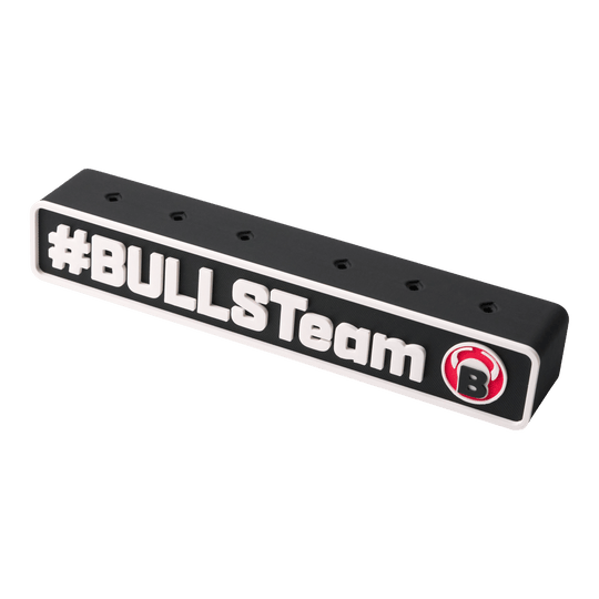 Das Bild zeigt einen schwarzen Darts-Halter mit der Aufschrift "#BULLSTeam" und einem roten Bullen-Logo. Der Halter hat mehrere Löcher zur Aufbewahrung von Dartpfeilen.