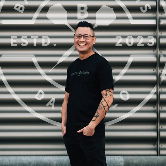 T-shirt „Barrels and Shafts” – czarny Ein Mann trägt ein schwarzes "Barrels and Shafts"-T-Shirt und steht vor einer metallenen Wand mit großem Logo. Er lächelt und hat markante Tattoos am rechten Arm.