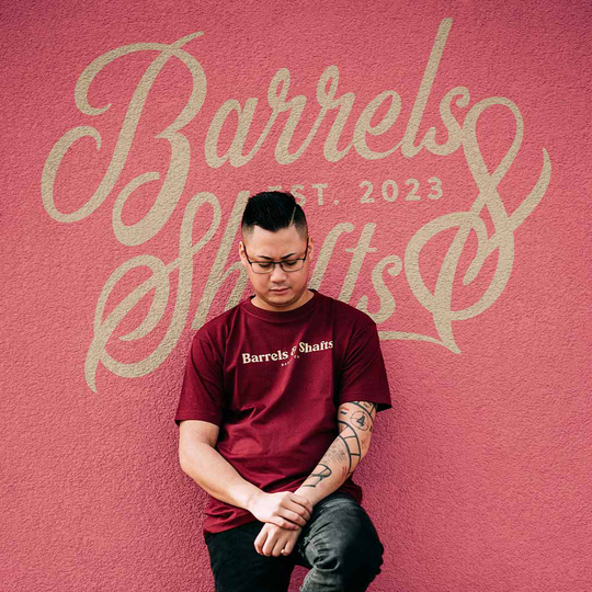 Ein Mann trägt ein bordeauxrotes T-Shirt mit der Aufschrift "Barrels & Shafts". Er steht vor einer Wand mit dem gleichen Schriftzug im Hintergrund.